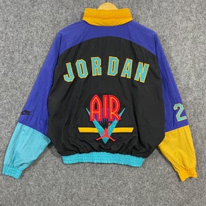 May include: A color-blocked windbreaker jacket featuring a black back panel with the text JORDAN and AIR in yellow and red. The jacket includes a red Jumpman logo, a yellow collar, blue sleeves, a teal cuff, a yellow sleeve end, and the number 2.