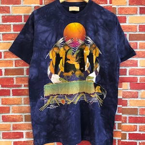 Puede incluir: Una camiseta azul marino tie-dye con una ilustración colorida de figuras nativas americanas y una puesta de sol. El texto "painted by The Mountain" está impreso en la parte inferior del diseño.