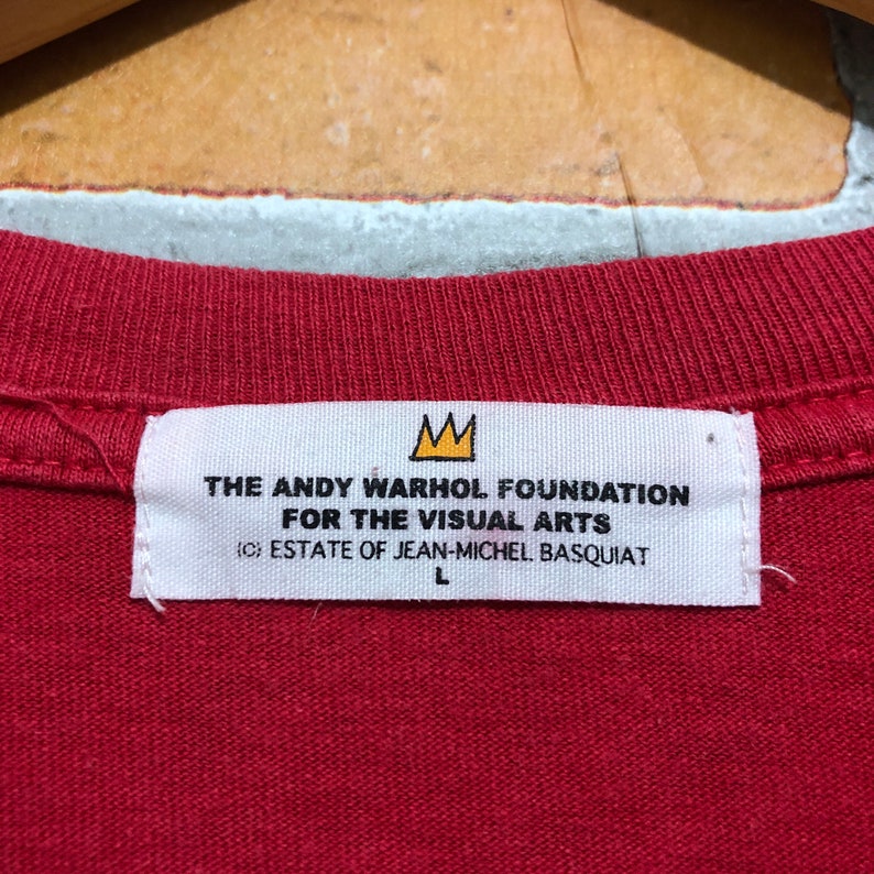 Puede incluir: Primer plano de una camiseta roja con una etiqueta blanca. La etiqueta muestra el texto "THE ANDY WARHOL FOUNDATION FOR THE VISUAL ARTS" y "(C) ESTATE OF JEAN-MICHEL BASQUIAT". Tambi&eacute;n se ve un logotipo de corona dorada.