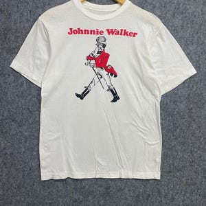 May include: White t-shirt featuring the Johnnie Walker logo in red, with a graphic of a man in a red coat and black boots. The man is walking with a cane. The shirt is hanging on a wooden hanger.