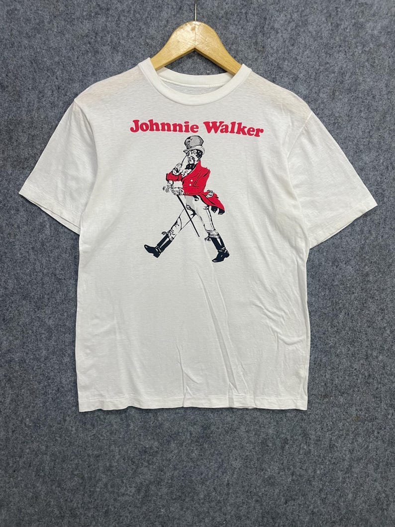 May include: White t-shirt featuring the Johnnie Walker logo in red, with a graphic of a man in a red coat and black boots. The man is walking with a cane. The shirt is hanging on a wooden hanger.
