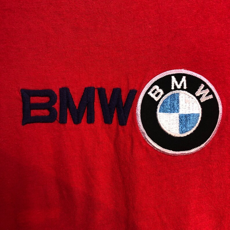 May include: Red fabric with the embroidered word "BMW" in navy blue and a circular BMW logo patch. The logo features a black border, white text, and a blue and white center.