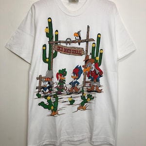 Peut inclure: T-shirt blanc avec un motif de dessin animé des personnages de Looney Tunes habillés en cow-boys. Le motif représente les personnages debout devant une pancarte qui indique "Daffy Duck's Corral".
