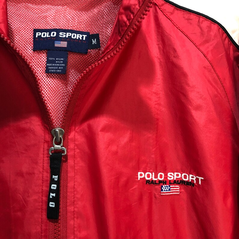 May include: Red nylon windbreaker jacket with a zipper closure and a Polo Sport Ralph Lauren logo embroidered on the front. The jacket has a mesh lining and a size tag that reads "M".