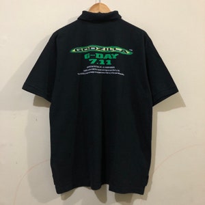 Può includere: Polo nero con una grafica verde e bianca di Godzilla G-Day 7.11 sul retro. La grafica include il testo "Godzilla" e "G-Day 7.11" con un design verde e bianco.