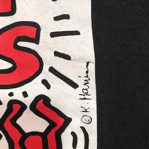 Puede incluir: Camiseta negra con un dise&ntilde;o gr&aacute;fico blanco con formas rojas y negras. El dise&ntilde;o incluye la firma del artista, "&copy; K. Haring". La camiseta tiene una est&eacute;tica sencilla y audaz.