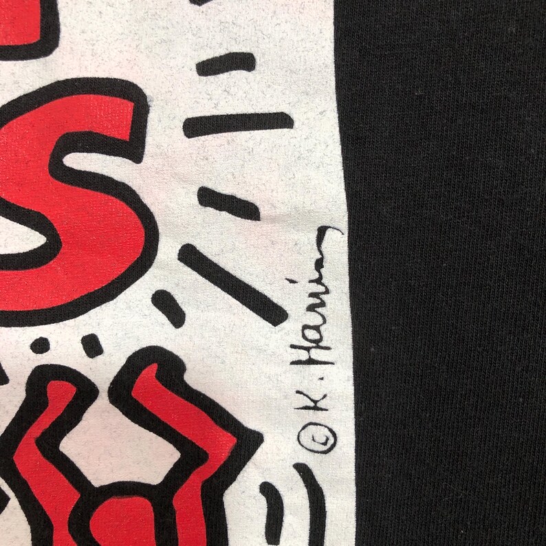 Puede incluir: Camiseta negra con un dise&ntilde;o gr&aacute;fico blanco con formas rojas y negras. El dise&ntilde;o incluye la firma del artista, "&copy; K. Haring". La camiseta tiene una est&eacute;tica sencilla y audaz.