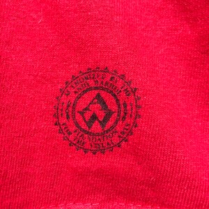 Puede incluir: Primer plano de una tela roja con un emblema circular negro. El emblema presenta una "A" y una "W" estilizadas dentro de un c&iacute;rculo, rodeadas de texto. La tela tiene una apariencia texturizada.