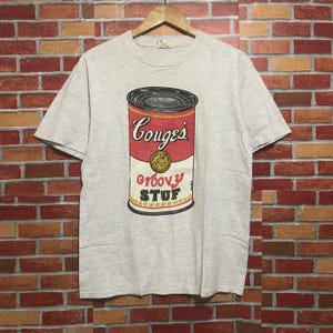 May include: A light gray t-shirt featuring a graphic of a soup can with the text "Gouges" and "Groovy STUF". The can design is red, white, and black with a gold emblem. The shirt has short sleeves and a crew neck.