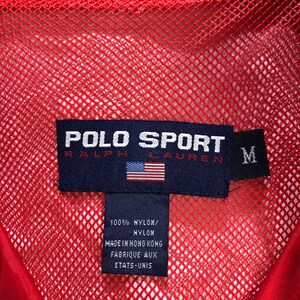 May include: A red mesh jacket with a black Polo Sport Ralph Lauren label. The label has the text "POLO SPORT RALPH LAUREN" and a small American flag. The label also has a size tag that says "M". The label has the text "100% NYLON/NYLON MADE IN HONG KONG FABRIQUE AUX ETATS-UNIS".
