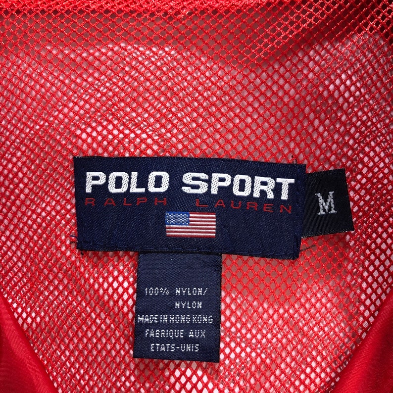 May include: A red mesh jacket with a black Polo Sport Ralph Lauren label. The label has the text "POLO SPORT RALPH LAUREN" and a small American flag. The label also has a size tag that says "M". The label has the text "100% NYLON/NYLON MADE IN HONG KONG FABRIQUE AUX ETATS-UNIS".