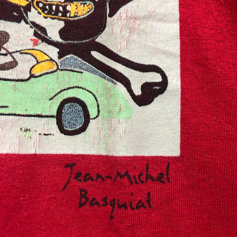 Puede incluir: Camiseta roja con un gr&aacute;fico de estilo pop art de un personaje de dibujos animados conduciendo un coche verde claro. El nombre del artista, "Jean-Michel Basquiat", est&aacute; escrito en escritura negra debajo de la imagen.