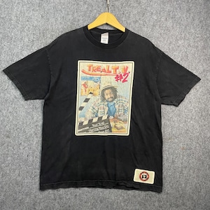 Puede incluir: Una camiseta negra de manga corta con un estampado gráfico de una persona con rastas que lleva una camisa de cuadros. El estampado incluye el texto "TREAL T.V #2" y una claqueta de cine. Un pequeño parche rectangular con un logo está abajo.
