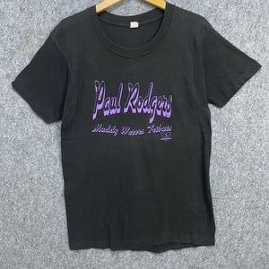 May include: Black t-shirt with the text "Paul Rodgers" in purple script above "Muddy Waters Tribute" and a logo. The shirt is hanging on a wooden hanger against a gray background. The t-shirt has a crew neck and short sleeves.