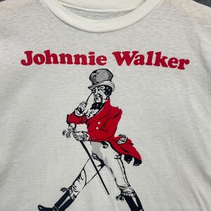 May include: White crew neck sweatshirt featuring the text "Johnnie Walker" in red above a graphic of a man in a red coat and black boots, walking with a cane. The graphic is in black and white.