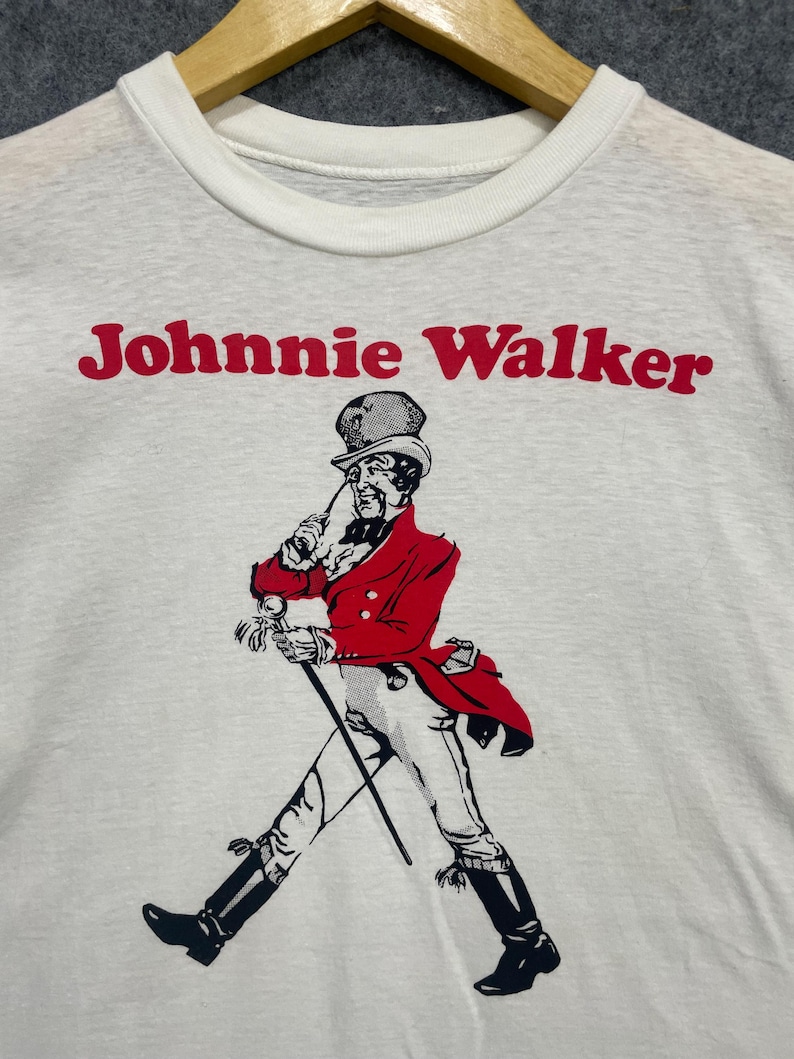 May include: White crew neck sweatshirt featuring the text "Johnnie Walker" in red above a graphic of a man in a red coat and black boots, walking with a cane. The graphic is in black and white.