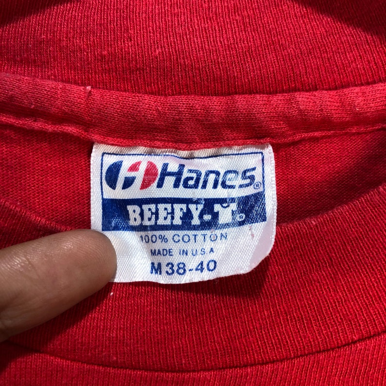 May include: Close-up of a red t-shirt with a Hanes BEEFY-T label. The label is white with blue and red text, stating "100% Cotton" and "M 38-40". The shirt is made in the USA.