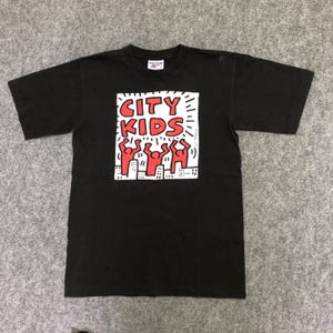 Puede incluir: Camiseta negra con un dise&ntilde;o gr&aacute;fico. El dise&ntilde;o presenta las palabras "CITY KIDS" en rojo, figuras rojas y edificios blancos. La camiseta tiene cuello redondo.