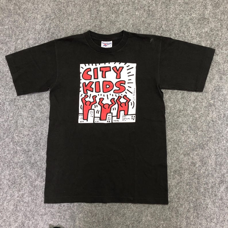 Puede incluir: Camiseta negra con un dise&ntilde;o gr&aacute;fico. El dise&ntilde;o presenta las palabras "CITY KIDS" en rojo, figuras rojas y edificios blancos. La camiseta tiene cuello redondo.