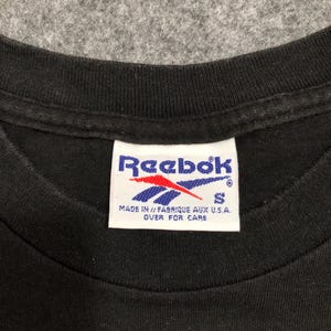 Puede incluir: Primer plano del cuello de una camiseta negra con una etiqueta Reebok. La etiqueta es blanca con el logotipo de Reebok en rojo y azul. La etiqueta tambi&eacute;n incluye el texto "MADE IN // FABRIQUE AUX U.S.A. OVER FOR CARE" y la talla "S".