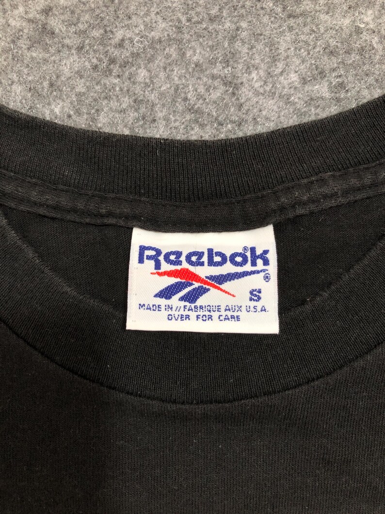 Puede incluir: Primer plano del cuello de una camiseta negra con una etiqueta Reebok. La etiqueta es blanca con el logotipo de Reebok en rojo y azul. La etiqueta tambi&eacute;n incluye el texto "MADE IN // FABRIQUE AUX U.S.A. OVER FOR CARE" y la talla "S".