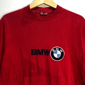 May include: Red t-shirt with the BMW logo embroidered on the front. The logo is a circular patch with the BMW emblem in the center. The text "BMW" is embroidered in navy blue.
