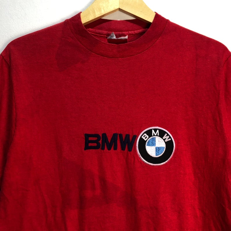 May include: Red t-shirt with the BMW logo embroidered on the front. The logo is a circular patch with the BMW emblem in the center. The text "BMW" is embroidered in navy blue.