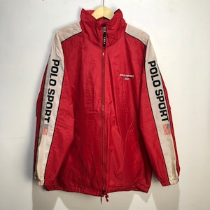 May include: Red windbreaker jacket with white and black stripes down the sleeves. The jacket has a zipper closure and a drawstring at the bottom. The text "POLO SPORT" is printed on the sleeves and the front of the jacket. The jacket has a small American flag on the sleeve.