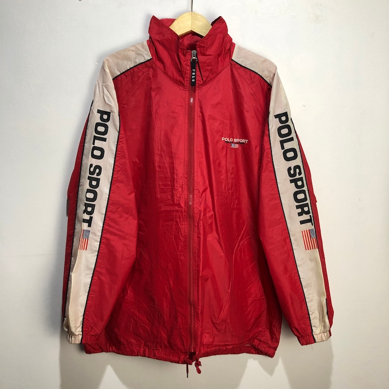 May include: Red windbreaker jacket with white and black stripes down the sleeves. The jacket has a zipper closure and a drawstring at the bottom. The text "POLO SPORT" is printed on the sleeves and the front of the jacket. The jacket has a small American flag on the sleeve.
