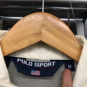 May include: Cream-colored Polo Sport Ralph Lauren jacket with a navy blue label featuring the brand name and the American flag. The jacket is size medium.