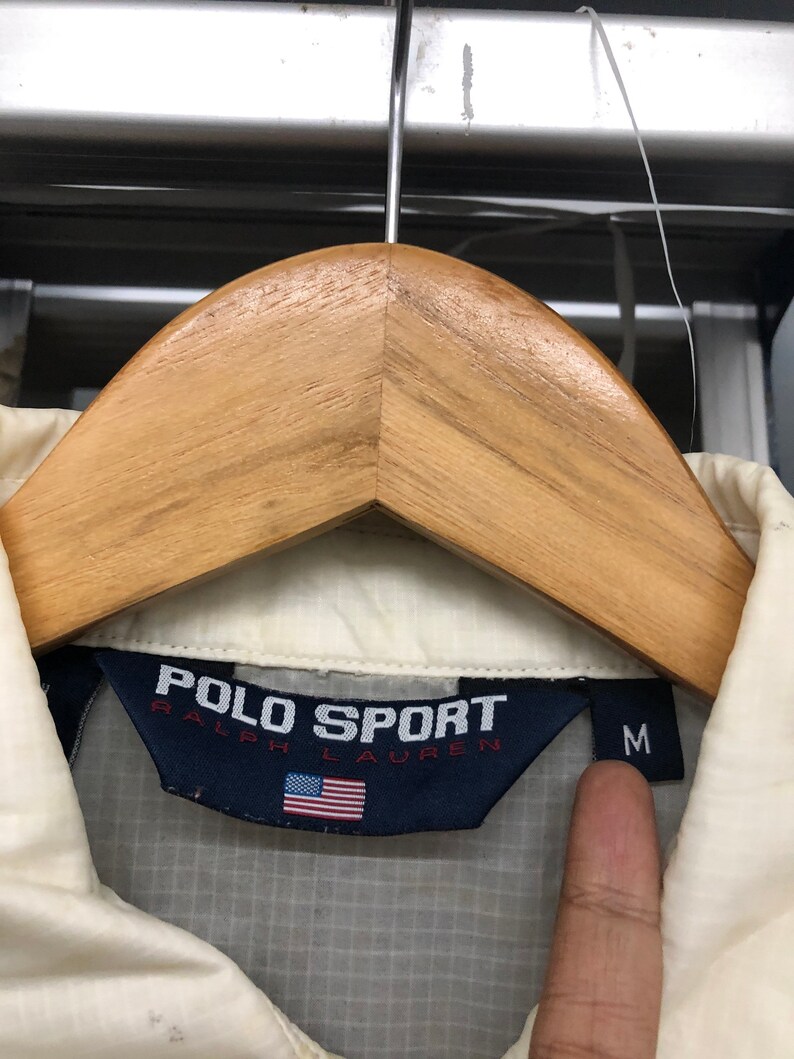 May include: Cream-colored Polo Sport Ralph Lauren jacket with a navy blue label featuring the brand name and the American flag. The jacket is size medium.