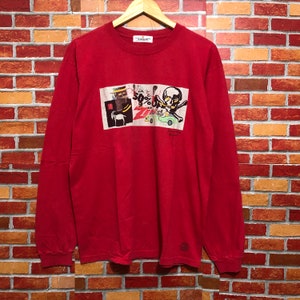 Puede incluir: Una camiseta roja de manga larga con un estampado gr&aacute;fico. El estampado presenta una calavera, un coche y texto que dice "50% OFF" y "Z". La camiseta est&aacute; colgada en una percha de madera.