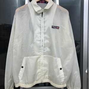 May include: Off-white, lightweight jacket with a full zipper closure and a small pocket on the front. The jacket features a collared neckline and a small patch with the word "POLO" and a flag design. The jacket is hanging on a wooden hanger.