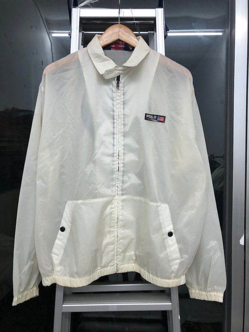 May include: Off-white, lightweight jacket with a full zipper closure and a small pocket on the front. The jacket features a collared neckline and a small patch with the word "POLO" and a flag design. The jacket is hanging on a wooden hanger.
