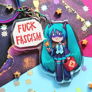 May include: An acrylic keychain featuring a cartoon character with long teal hair, holding a lit match and a red fuel canister. A white speech bubble attached to the character displays the red text FUCK FASCISM. The keychain has a rainbow metal ring.