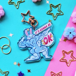 May include: A blue and pink acrylic charm featuring a cartoon bunny with muscles, holding a pink heart with the text "It's gonna be OK". The charm is on a pink and blue background with gold stars and other decorative elements.