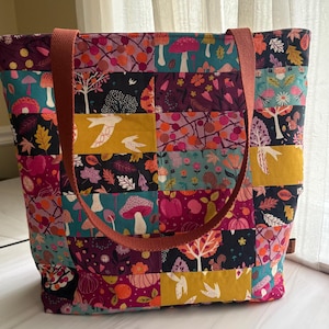 May include: A patchwork tote bag featuring a colorful print of mushrooms, squirrels, birds, leaves, and flowers in shades of teal, mustard yellow, magenta, and dark navy. The bag has two sturdy, solid-colored terracotta fabric straps.