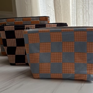 May include: Three zippered pouches with a checkerboard pattern. The pouches feature a mix of colors, including orange, black, and gray. The pouches are made of fabric and have a rectangular shape. The zippers are black.