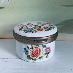May include: A white porcelain trinket box with a floral design. The lid is closed and has a brass rim. The box is decorated with a floral pattern in shades of pink, red, blue, and green.