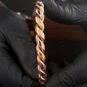 Può includere: Un bracciale in metallo color rame caratterizzato da un design a corda intrecciata continua. Il bracciale presenta una superficie lucida e riflettente con solchi profondi che seguono il motivo a spirale lungo l'intera fascia circolare.