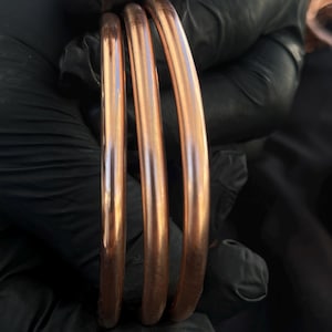 May include: Three smooth, polished copper bangles with a rounded profile. The bracelets are metallic, reddish-brown in color, and feature a simple, continuous circular design without any additional ornamentation, clasps, or surface textures.