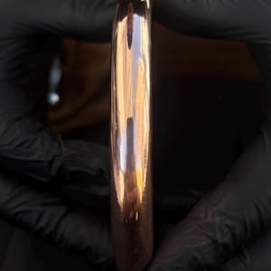 May include: A smooth, polished copper-colored bangle bracelet with a rounded, seamless profile. The metal surface is highly reflective, showing light highlights along its curved edge, presenting a solid, circular form without any visible clasps or markings.