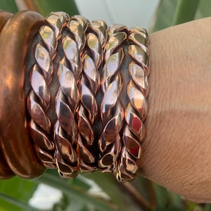 Puede incluir: Un conjunto de varias pulseras de cobre pulido con un diseño de cuerda trenzada. Las pulseras tienen un acabado metálico reflectante y se llevan juntas en la muñeca, mostrando un patrón de espiral repetitivo en cada banda individual.