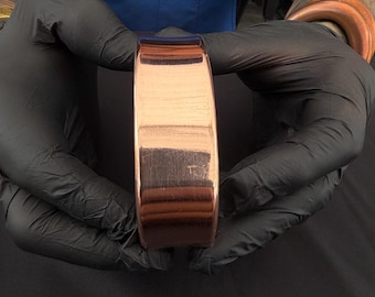 May include: A thick, polished copper cuff bracelet with a smooth, reflective metallic surface. The band features a wide, flat profile with rounded edges, showcasing the warm, reddish-brown hue of the metal in a simple, unadorned design.