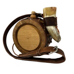 Puede incluir: Una cantina de madera envuelta en cuero marrón con una taza para beber de cuerno unida. La cantina tiene una correa para el hombro y una hebilla de metal.