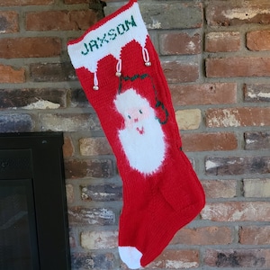 May include: A red knitted Christmas stocking with a white Santa Claus face and the name "JAXSON" in white letters. The stocking hangs on a brick fireplace, with small bells and white trim.
