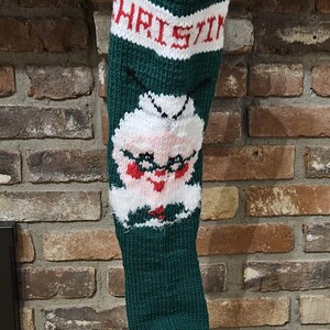 May include: A green knitted Christmas stocking with a white and red "CHRISTMAS" banner at the top and a white knitted face with red lips and glasses in the middle.