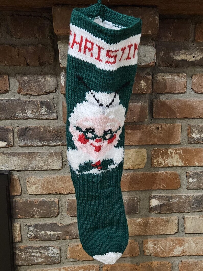 May include: A green knitted Christmas stocking with a white and red "CHRISTMAS" banner at the top and a white knitted face with red lips and glasses in the middle.