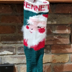 May include: A green, red, and white knitted Christmas stocking with a Santa Claus face and the name "Jenna" embroidered on the top.