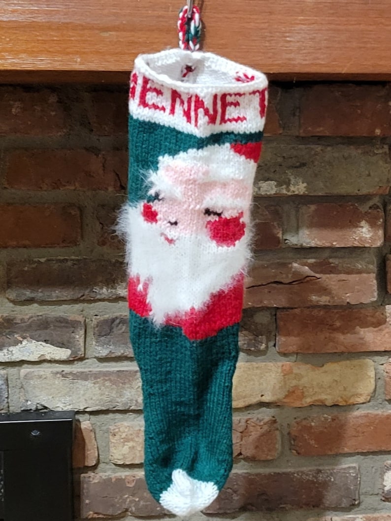 May include: A green, red, and white knitted Christmas stocking with a Santa Claus face and the name "Jenna" embroidered on the top.